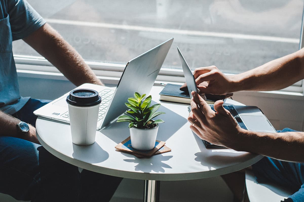 Due persone sedute a un tavolino con i propri laptop; sul tavolo sono presenti una pianta, un caffè da asporto e un'agenda